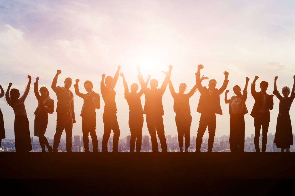 The image shows a group of people standing side by side, silhouetted against a bright, glowing background with the sun shining. The individuals have their arms raised in celebration or triumph, conveying a sense of unity, achievement, and joy. The background suggests a cityscape, adding to the atmosphere of success and accomplishment.