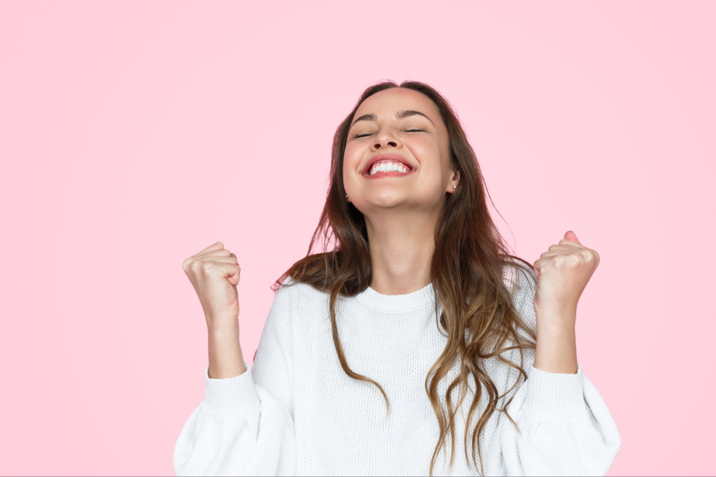 The image shows a young woman with long hair, wearing a white sweater, against a pink background. She has a joyful expression on her face, eyes closed, and is smiling broadly. Her fists are clenched in a celebratory gesture, conveying a sense of triumph, happiness, and accomplishment.